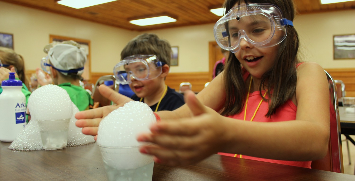 Young girl and boy with science safety goggles on, bubbling science experiement in front of them.