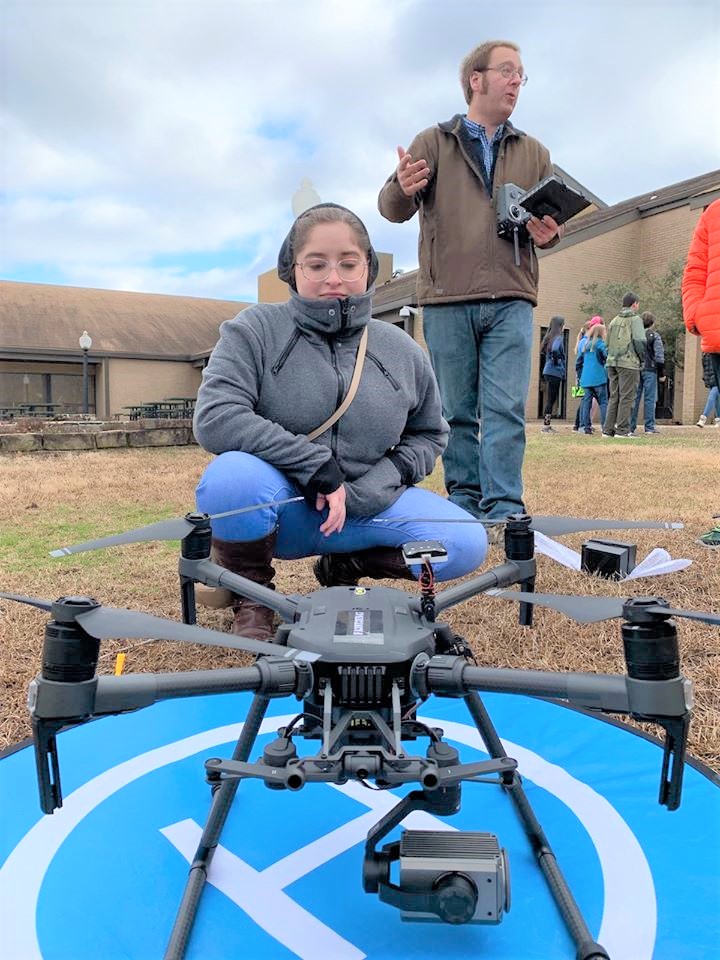 Female student kneeled down in front of a drone launch pad with drone on it. Other students and instructor in the background.