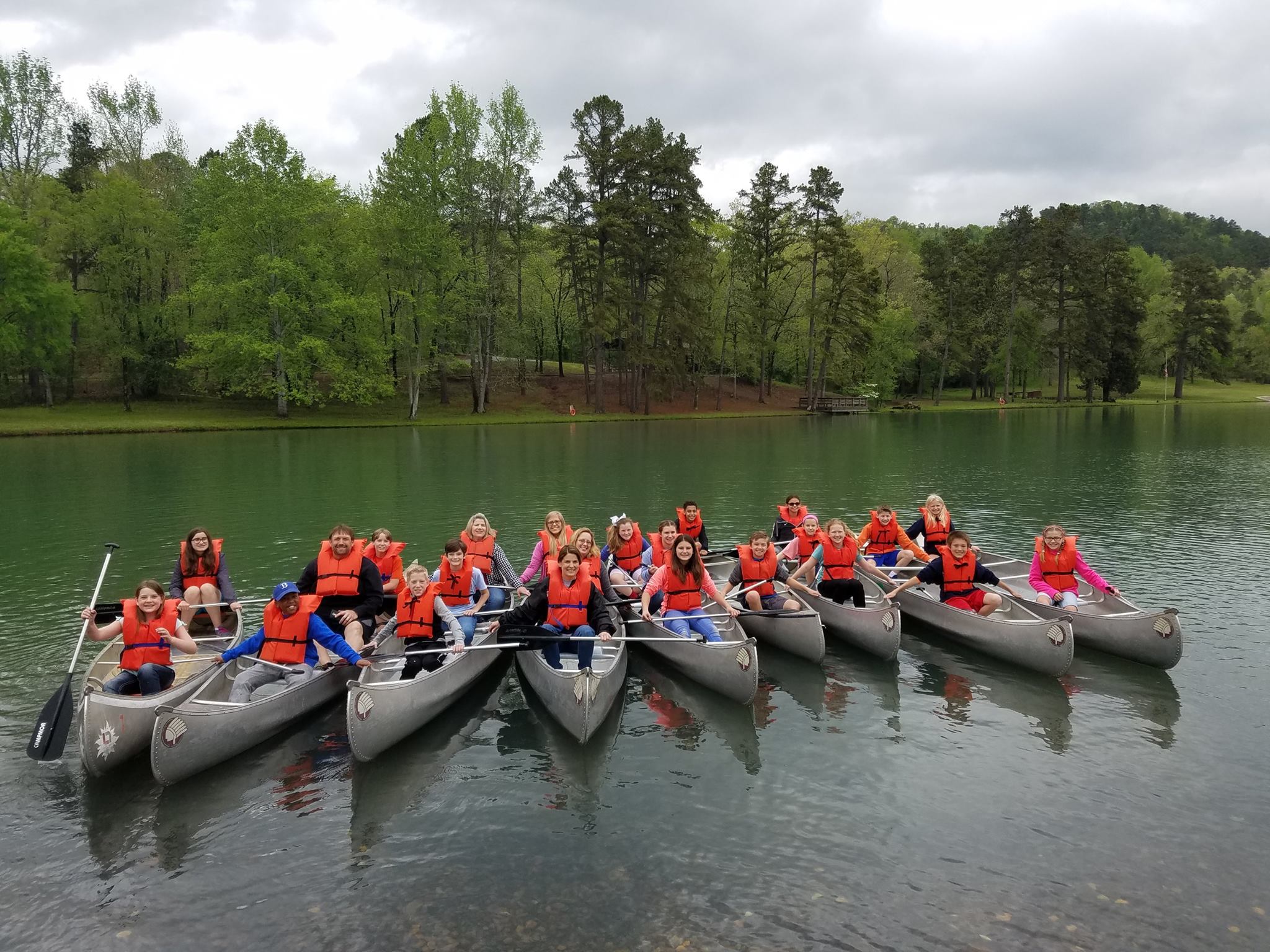 Large group of youth in multiple canoes, all wearing orange life vests, lake and trees in background
