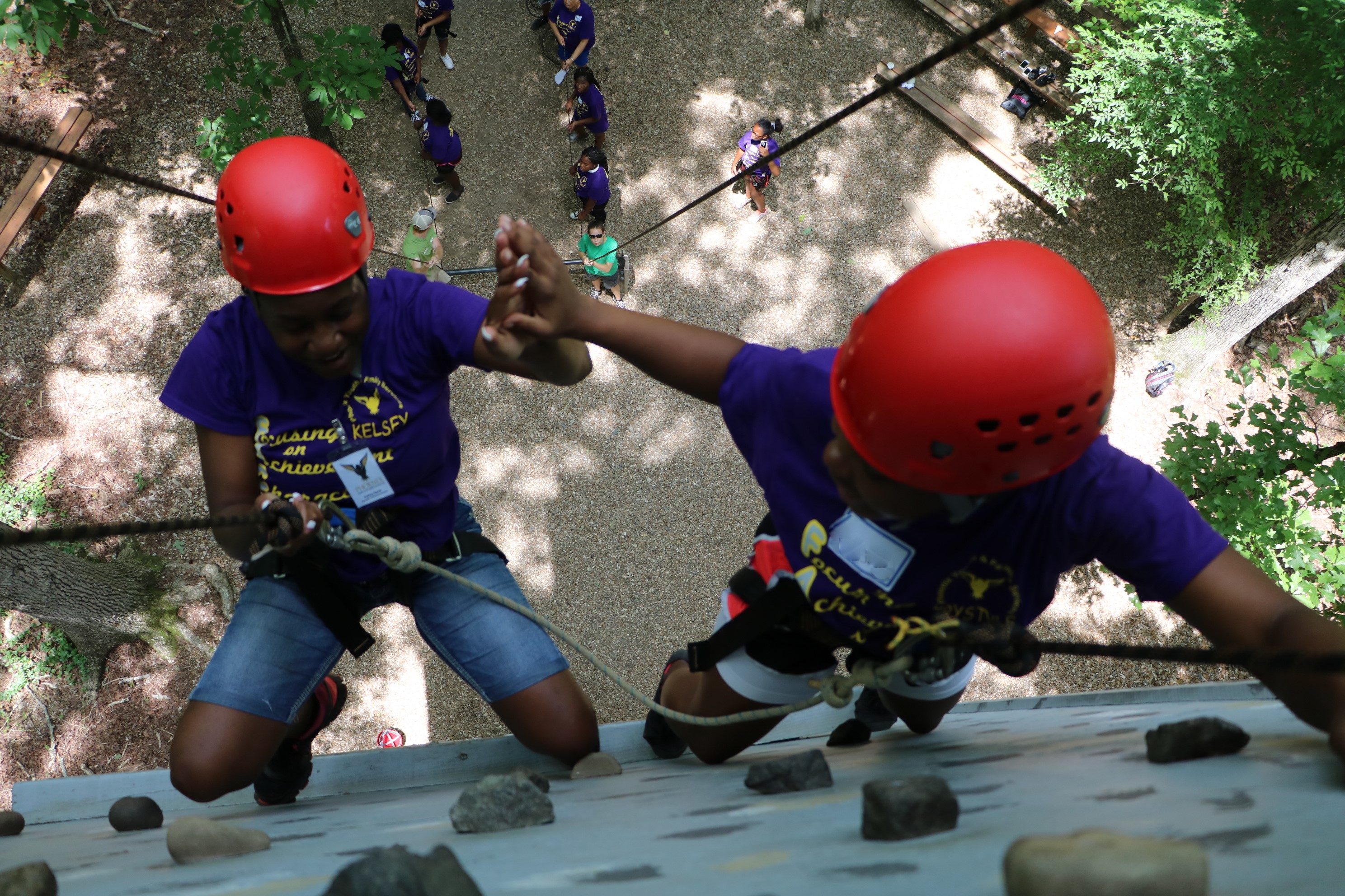 Looking down from the top of the rock wall structure at 2 youth in orange helmets climbing to the top. Youth are high-fiving.