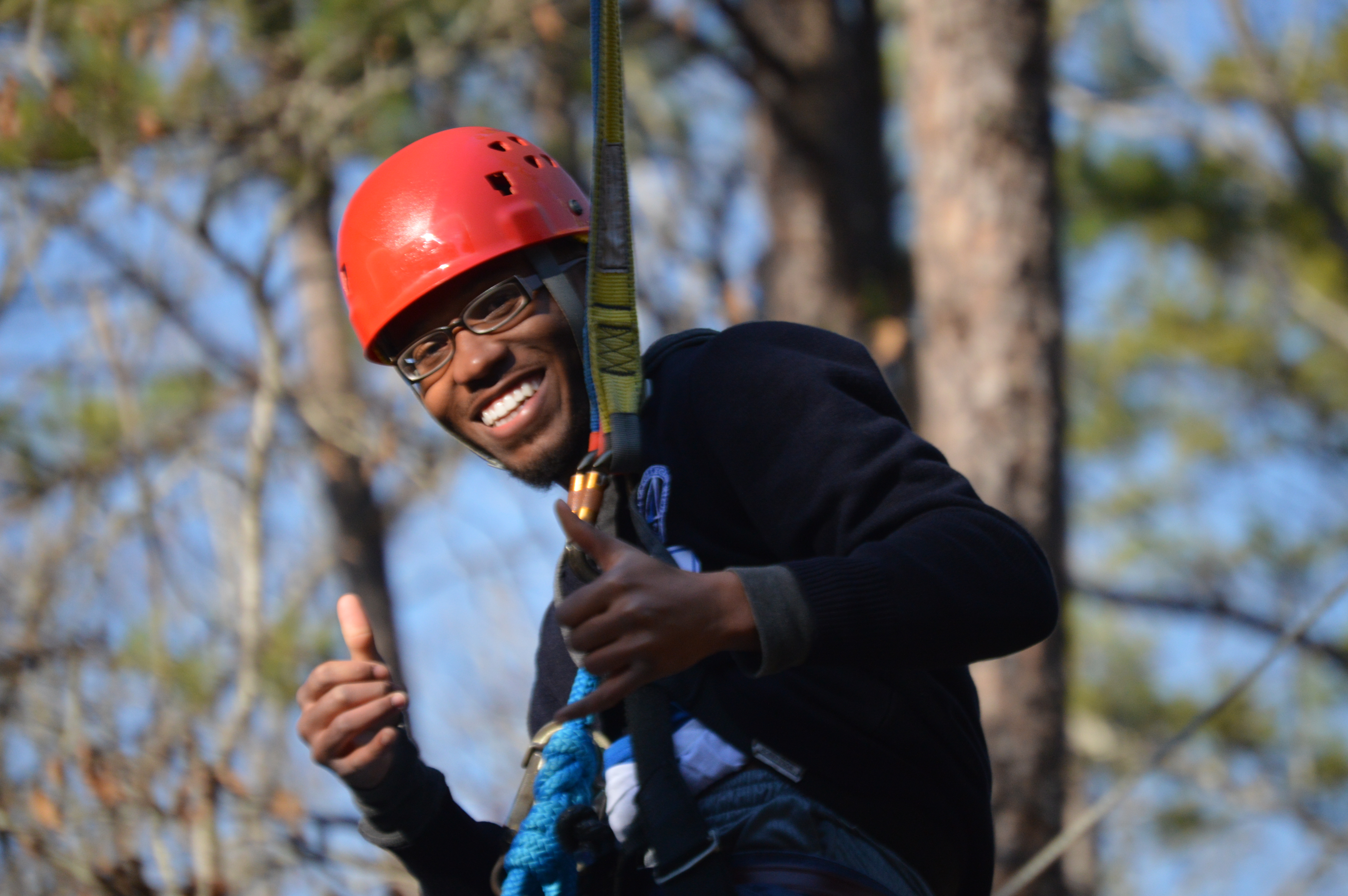Young male in harness and orange helmet, with 2 thumbs up, coming down the zip line. 