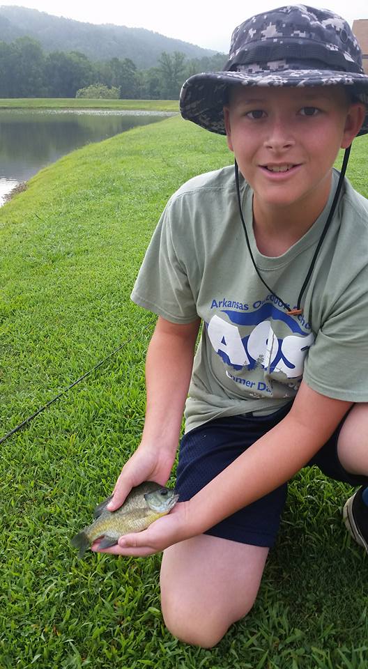 10 year old boy in AOS t-shirt kneeling next to a lake while holding a small fish.