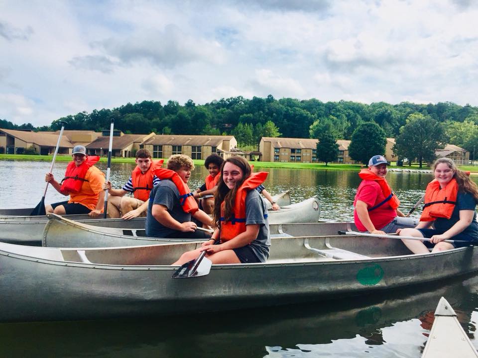 6 4-H youth in canoes, 2 per canoe, wearing orange life vests. Lake and 4-H Center buildings in background